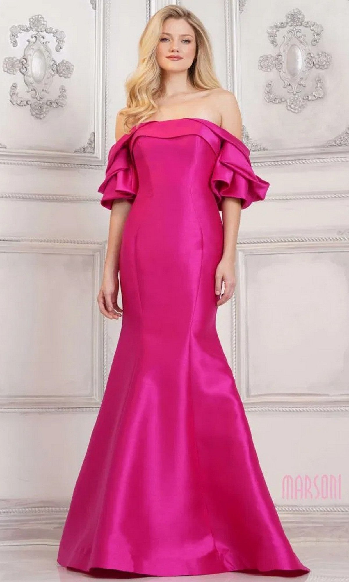 A woman poses against an ornate light wall in the Marsoni MV1290, a fuchsia long formal dress with off-the-shoulder ruffled sleeves and a mermaid silhouette.